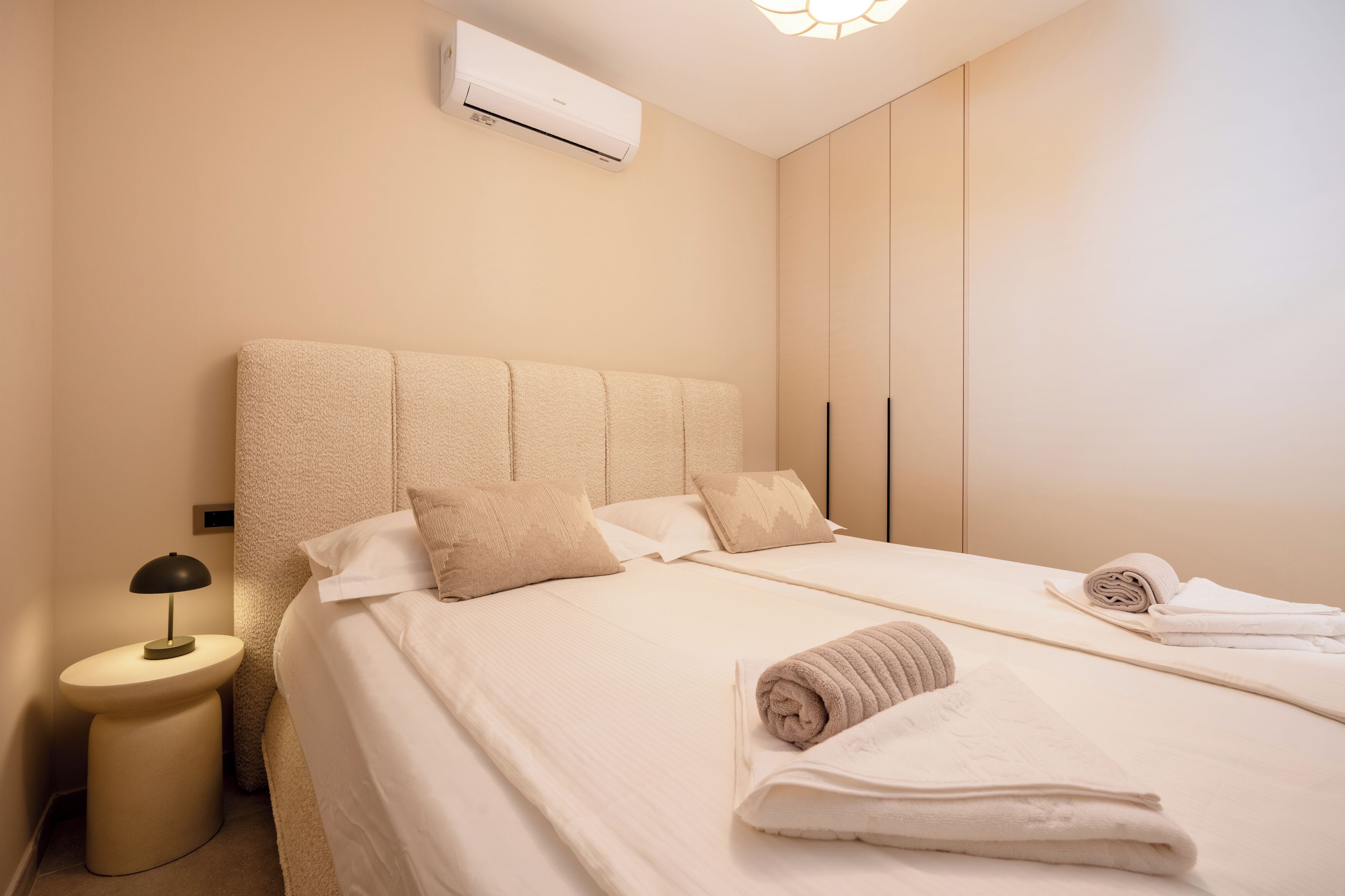 Ein ordentlich bezogenes Bett mit Kissen, eine Nachttischlampe und aufgerollte Handtücher in einem sauberen, hellen Zimmer mit Wand-Klimaanlage.