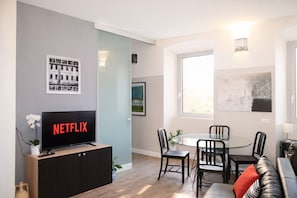 Ein modernes Wohnzimmer mit Flachbildfernseher, einem Esstisch mit Stühlen, einer Couch und einem großen Fenster.