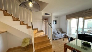 Ein modernes Interieur mit einer Holztreppe, einem grauen Sofa, einem Esstisch mit Stühlen und einem großen Fenster.