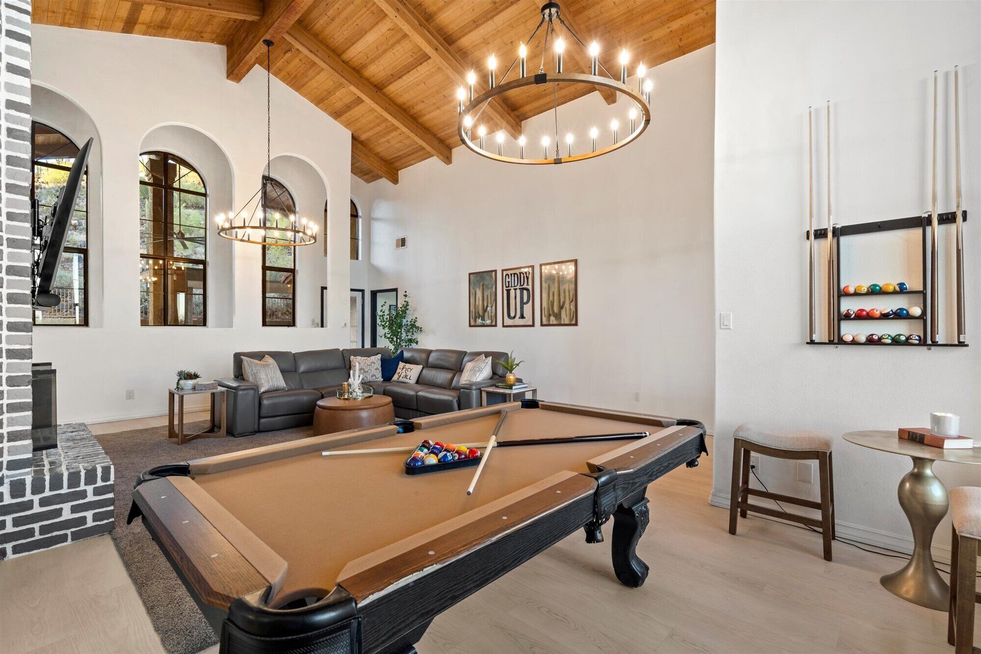 A pool table with billiard balls, a sofa set, and a pool cue rack in a room with wooden beams and a chandelier.