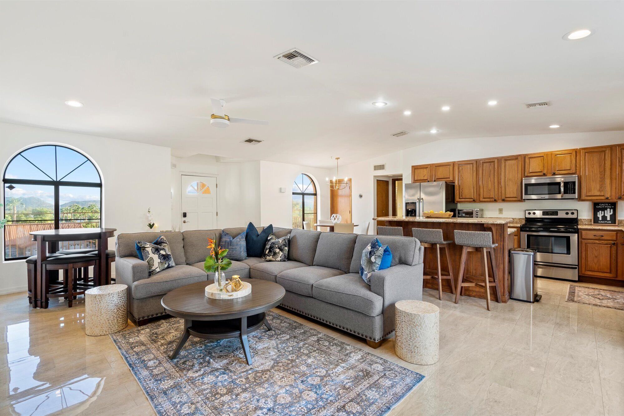 A spacious living room with a gray sectional sofa, a coffee table, and a patterned rug. A kitchen with wooden cabinets and stainless steel appliances is visible in the background.