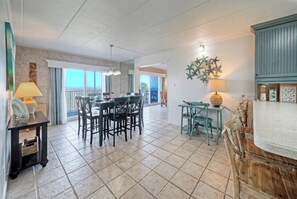 A spacious dining area with a large table and chairs, a small side table with a lamp, and a view to the outside. - 2-Min Walk to Beach + Boardwalk + Golf Cart Zone | Serenity by the Sea (Port Aransas)