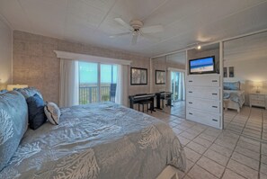 A bedroom with a bed, a desk, a piano, a TV, and a sliding door leading to another room. - 2-Min Walk to Beach + Boardwalk + Golf Cart Zone | Serenity by the Sea (Port Aransas)