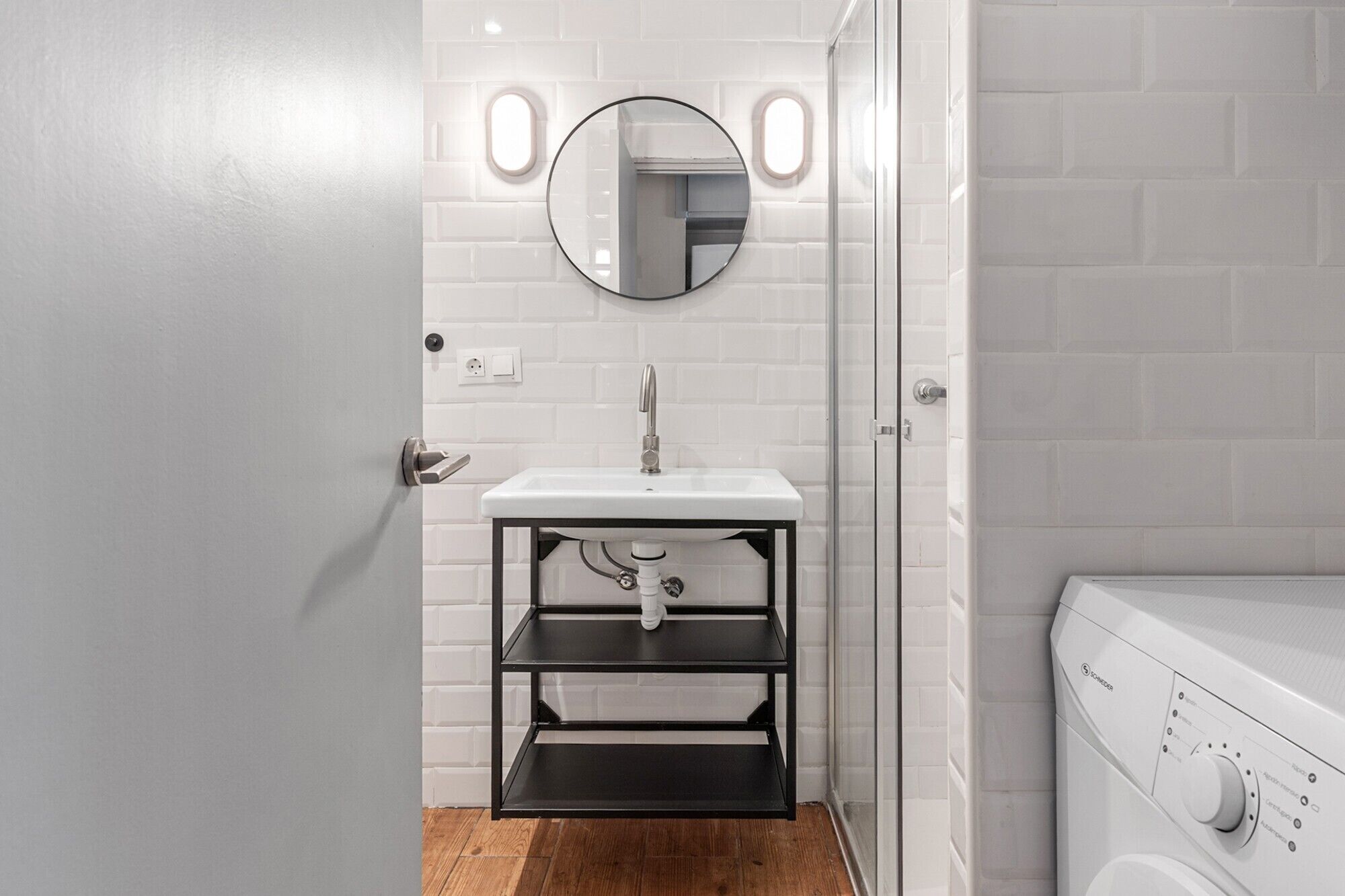 Un baño con un lavabo blanco, un espejo, una repisa metálica negra y una lavadora.