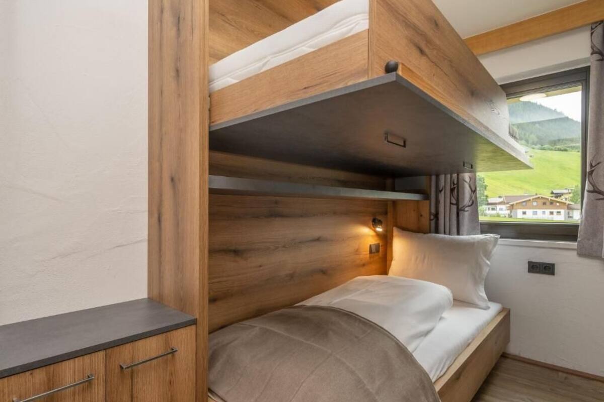 A bunk bed with a wooden headboard, a bedside table, and a window with a view of a house and hills.