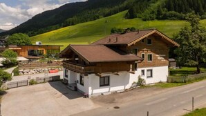 A two-story wooden house with a balcony, surrounded by a paved driveway and a grassy area. - Hofers Herzstück (Hinterglemm)