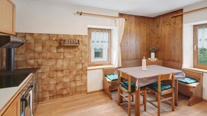 Een keuken met een fornuis, oven en raam met gordijnen. Er staat een eettafel met stoelen en een bankje.