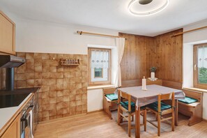 Een keuken met een fornuis, oven en houten kasten. Er is een eettafel met stoelen en een bank.