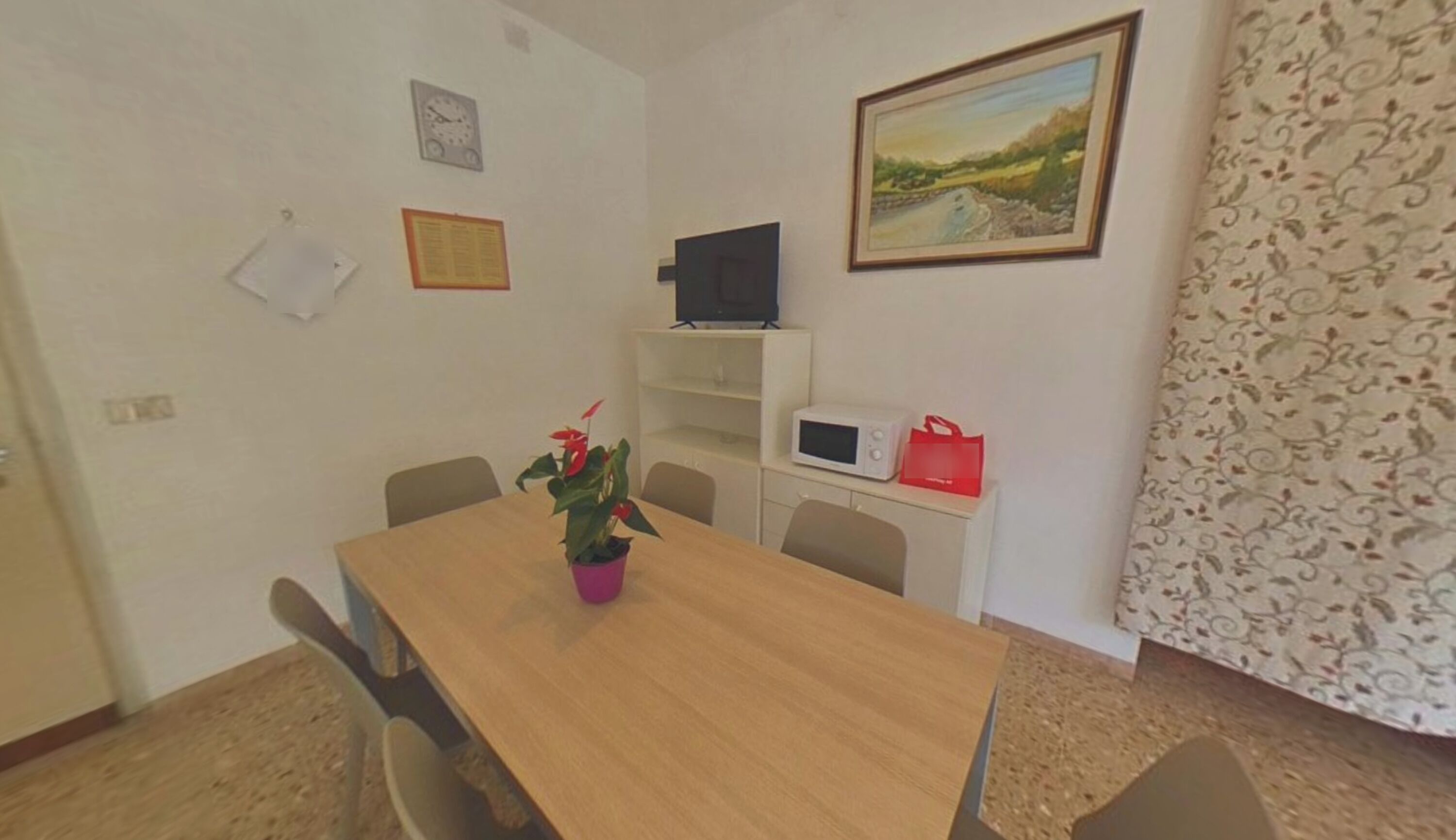 A room with a wooden table, chairs, a potted plant, a TV on a shelf, a microwave, and a painting on the wall.