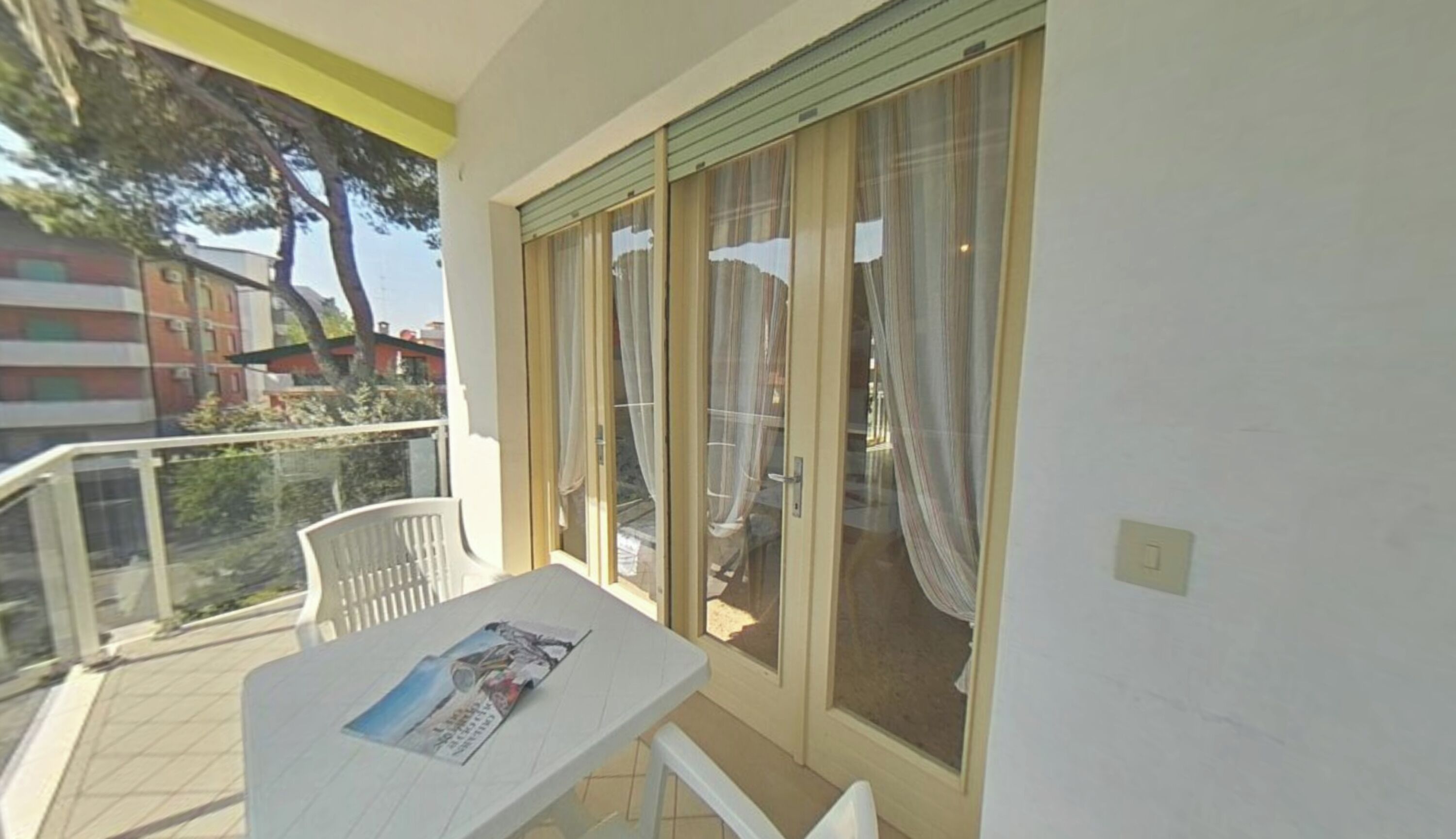 A balcony with a table and chairs, a window with curtains, and a view of buildings and trees.