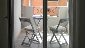Un balcon ouvert avec deux chaises et une table, une vue sur un immeuble avec un toit en tuiles, et une fenêtre avec des volets fermés.