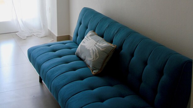 Un canapé de couleur bleu-vert avec un coussin à motifs, un climatiseur fixé au mur et deux tableaux représentant des scènes maritimes.