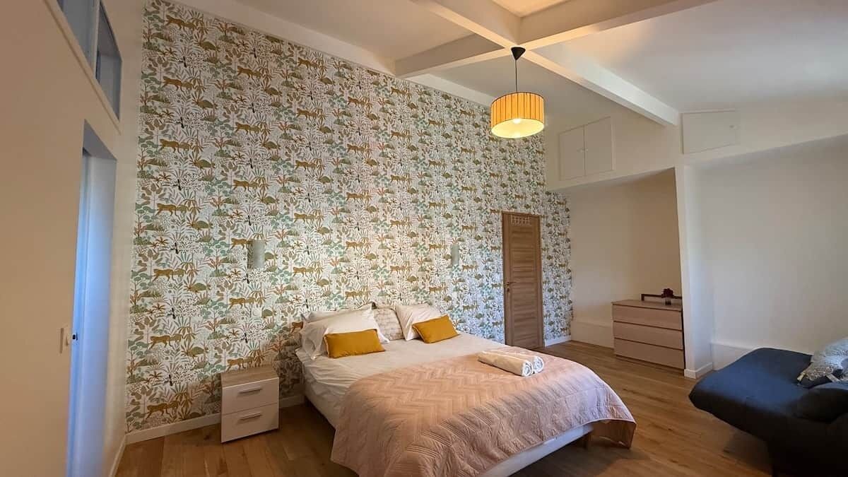 Un dormitorio con papel tapiz estampado, una cama con almohadas, una mesita de noche, un aparador y una lámpara colgante.