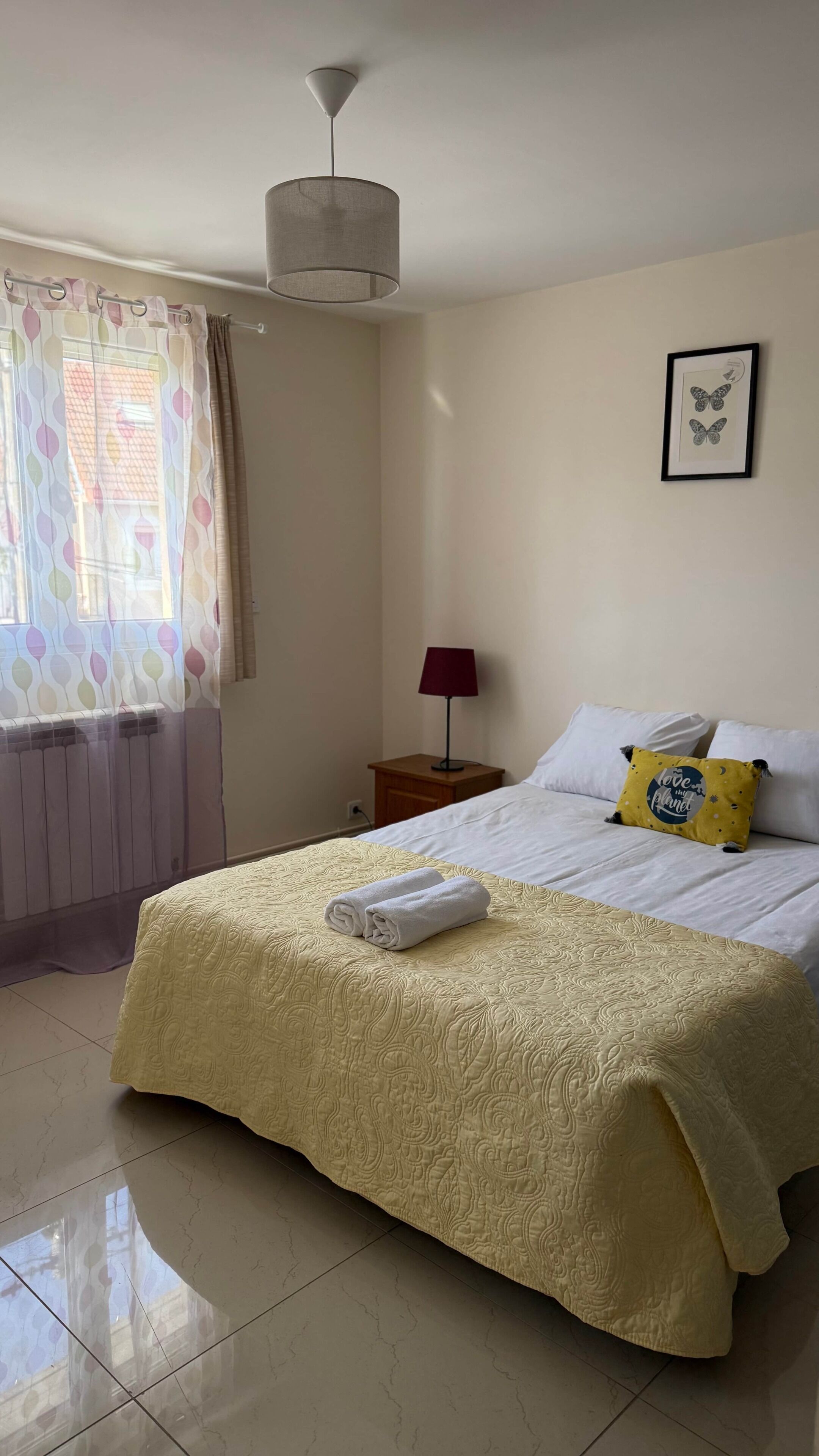 A neatly made bed with a yellow bedspread, a red bedside lamp, a framed butterfly picture, and a window with patterned curtains.