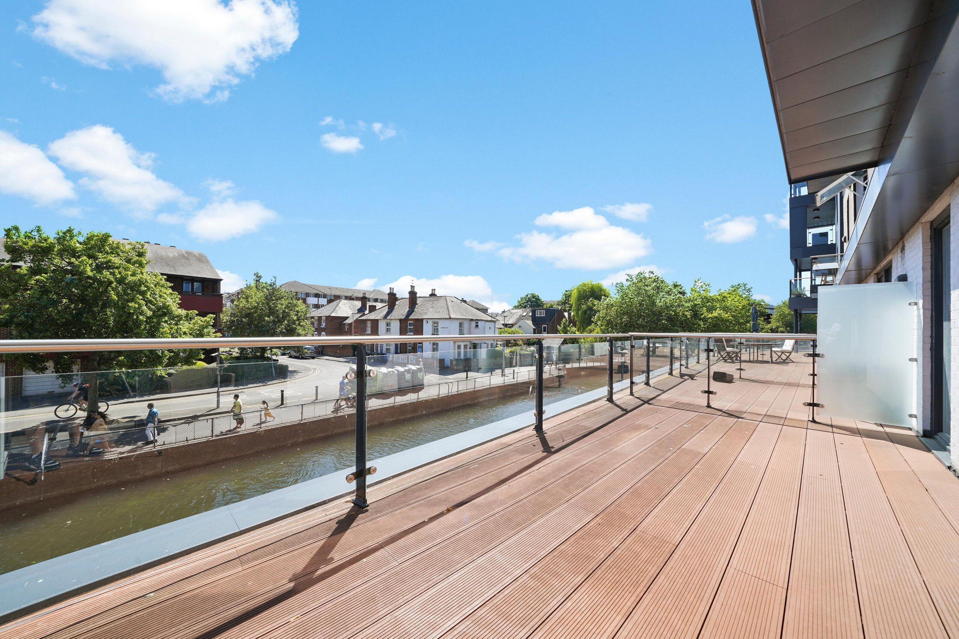 A wooden deck with a railing overlooking a canal, with buildings and trees in the background.
