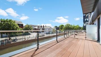 A wooden deck with a railing overlooking a canal, with buildings and trees in the background.