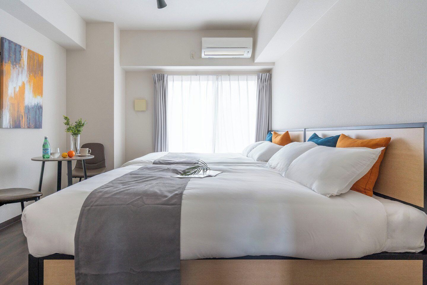A hotel room with a bed, a small table with a chair, a window with curtains, and an air conditioning unit.