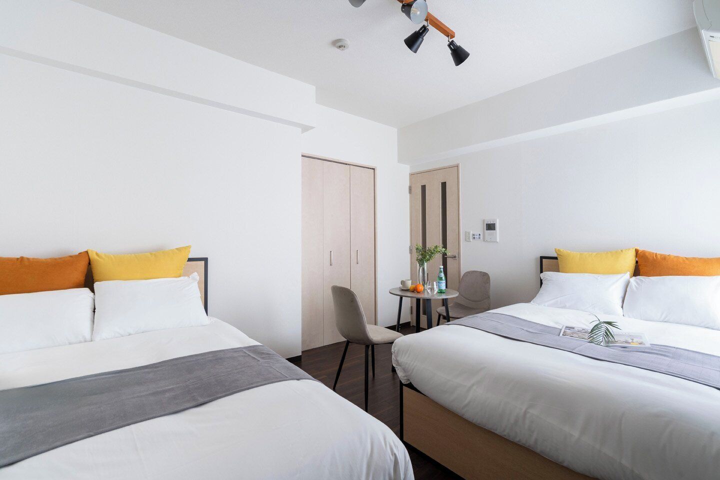 Une chambre d’hôtel avec deux lits, une petite table avec une chaise, une porte et un luminaire au plafond.