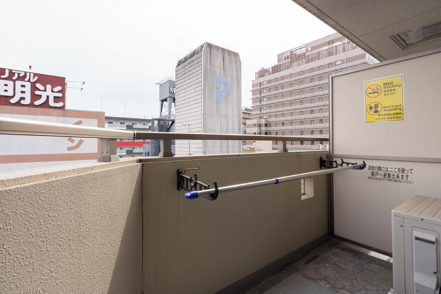 A balcony with a metal railing, a wall-mounted sign with a no-smoking and no-putting-anything-down warning, and a view of surrounding buildings.