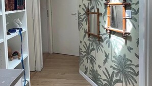 En hall med trägolv, en vit bokhylla med böcker och dekorativa föremål, och en vägg med ett tropiskt växtmönster.