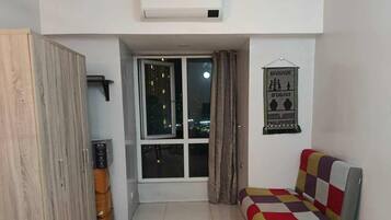 A living room with a sofa, a table, a wardrobe, a window with curtains, and a wall-mounted air conditioner.