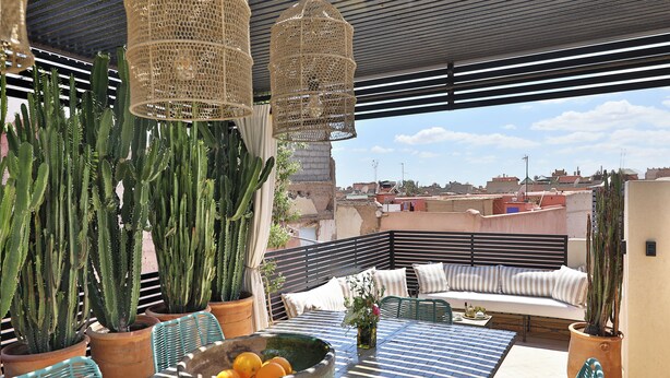 A rooftop terrace with a dining table, chairs, a sofa, potted cacti, and hanging wicker lamps. - Riad Della (Marrakech)