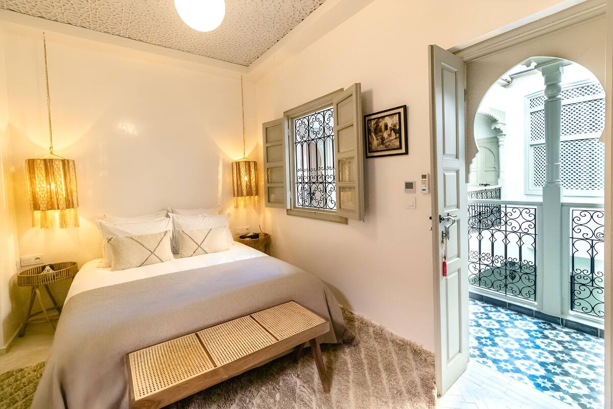 Quarto com cama, abajures, porta que dá para uma varanda com piso estampado.
