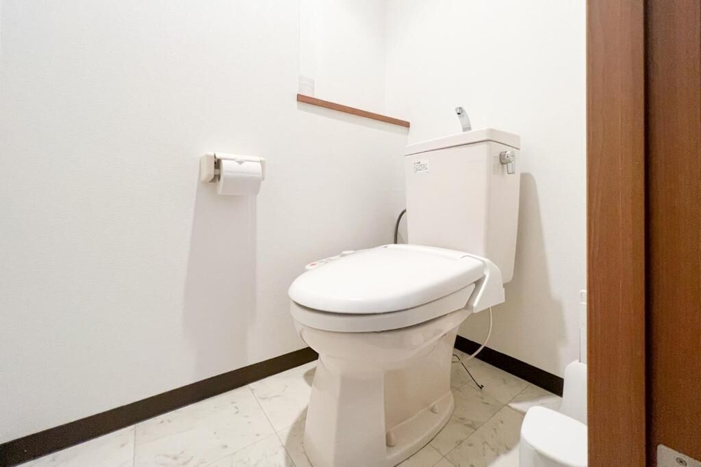 A white toilet with a tank, a roll of toilet paper on the wall, and a wooden shelf above the toilet.