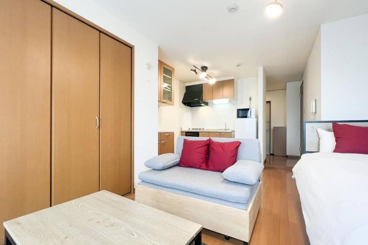 A compact room with a sofa, a small table, a wardrobe, and a kitchenette in the background.