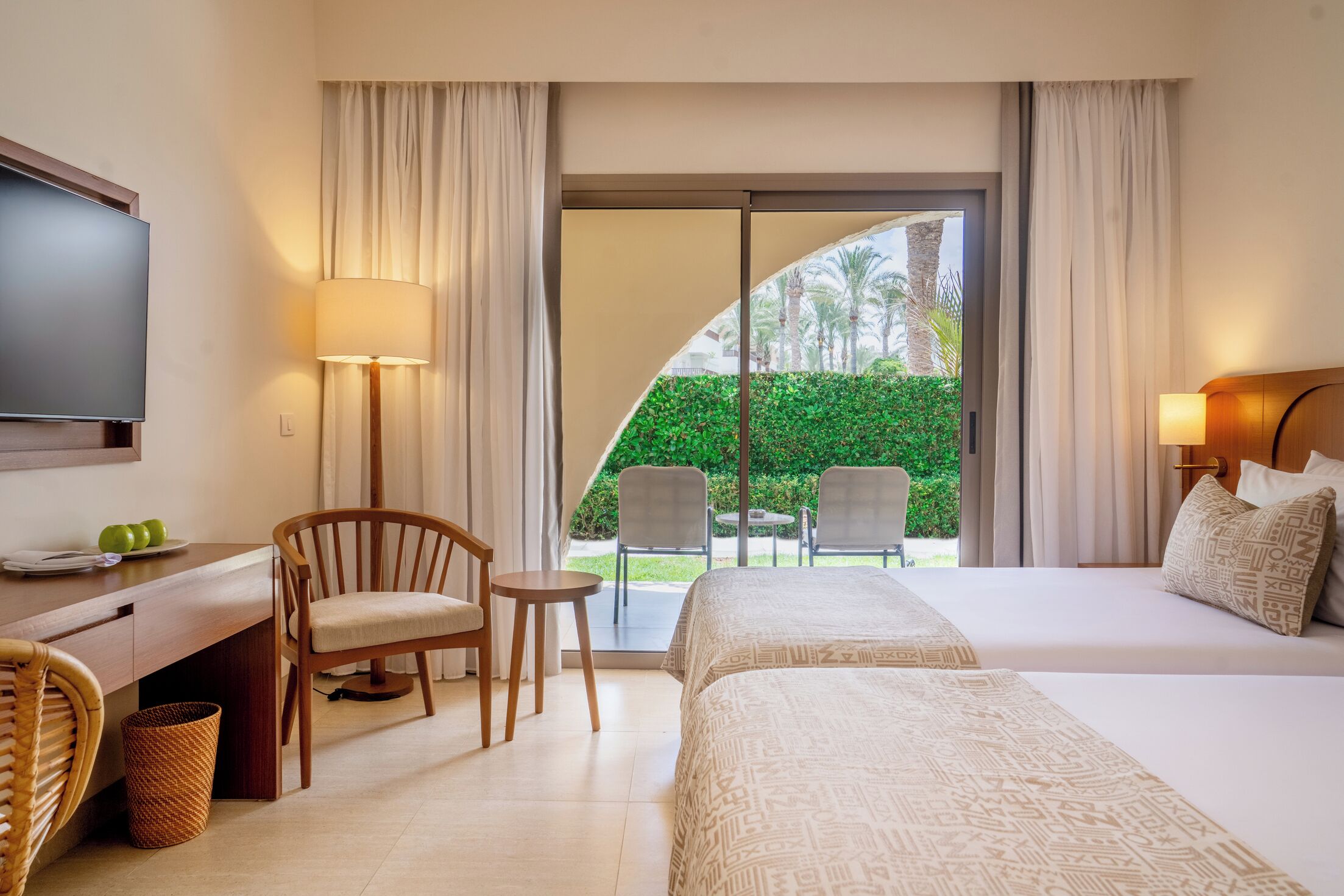 Habitación de hotel con cama, escritorio con televisor, silla, mesa pequeña y vista a un jardín por una puerta corredera.