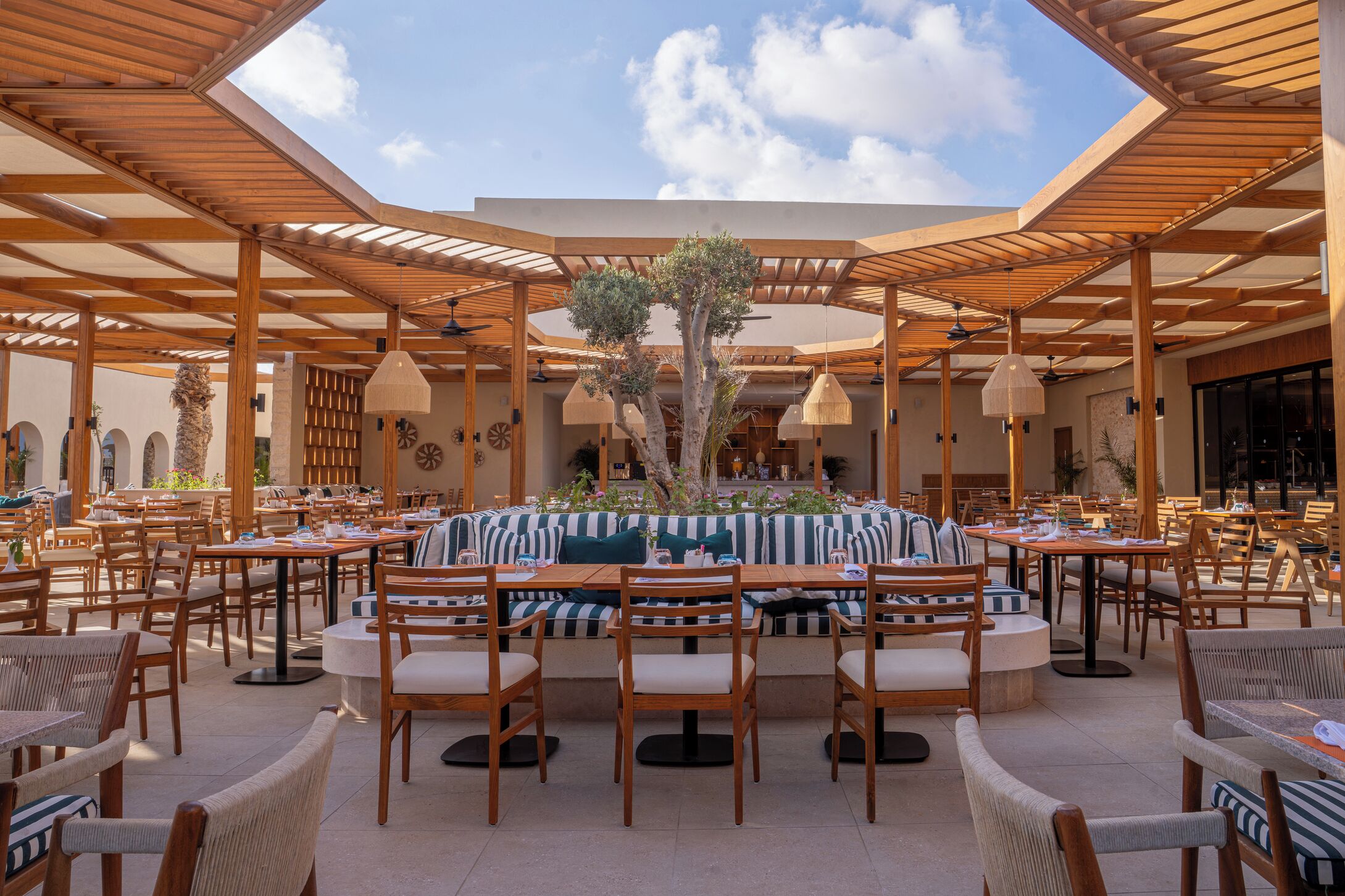 Un espace de restauration en plein air avec des tables et des chaises en bois, un coin salon central agrémenté d’une nappe à rayures et une pergola au-dessus.