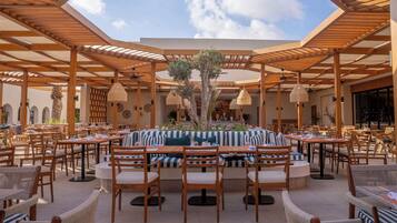 Un espace de restauration en plein air avec des tables et des chaises en bois, un coin salon central agrémenté d’une nappe à rayures et une pergola au-dessus.