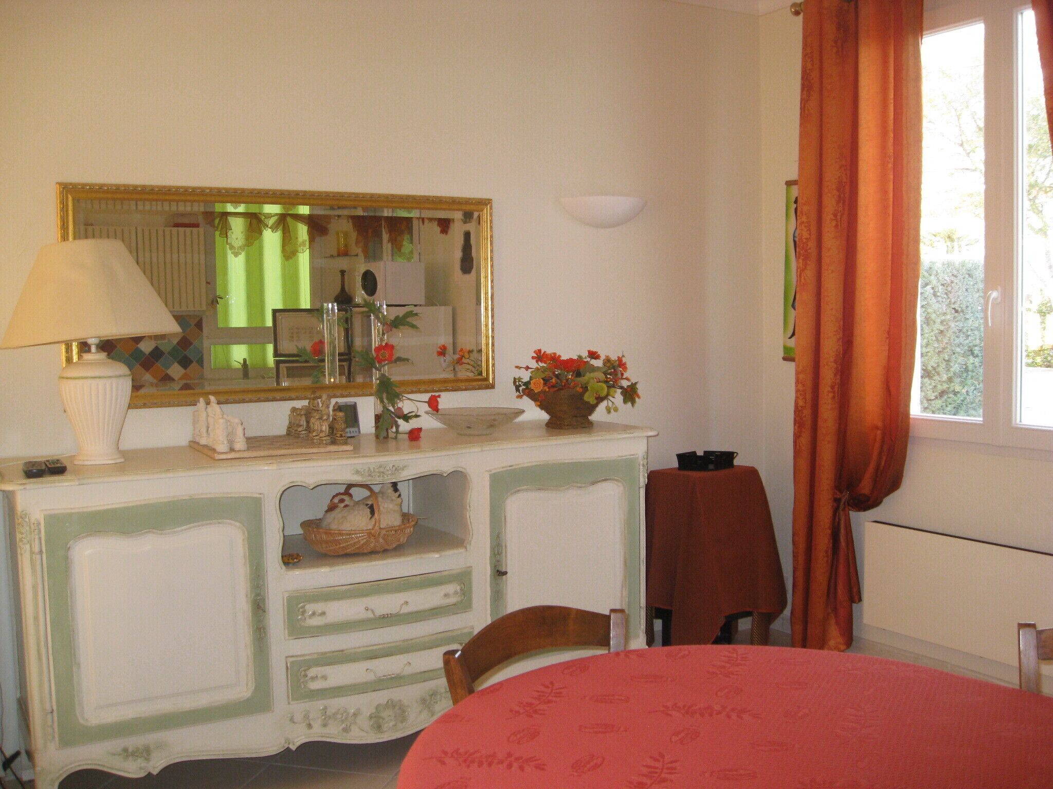 A room with a dresser, a mirror, a table with a red tablecloth, and a window with curtains.