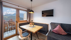 Ein Zimmer mit Holztisch, einem Sessel, einer Couch, einem Fernseher und einem Balkon mit Bergblick.