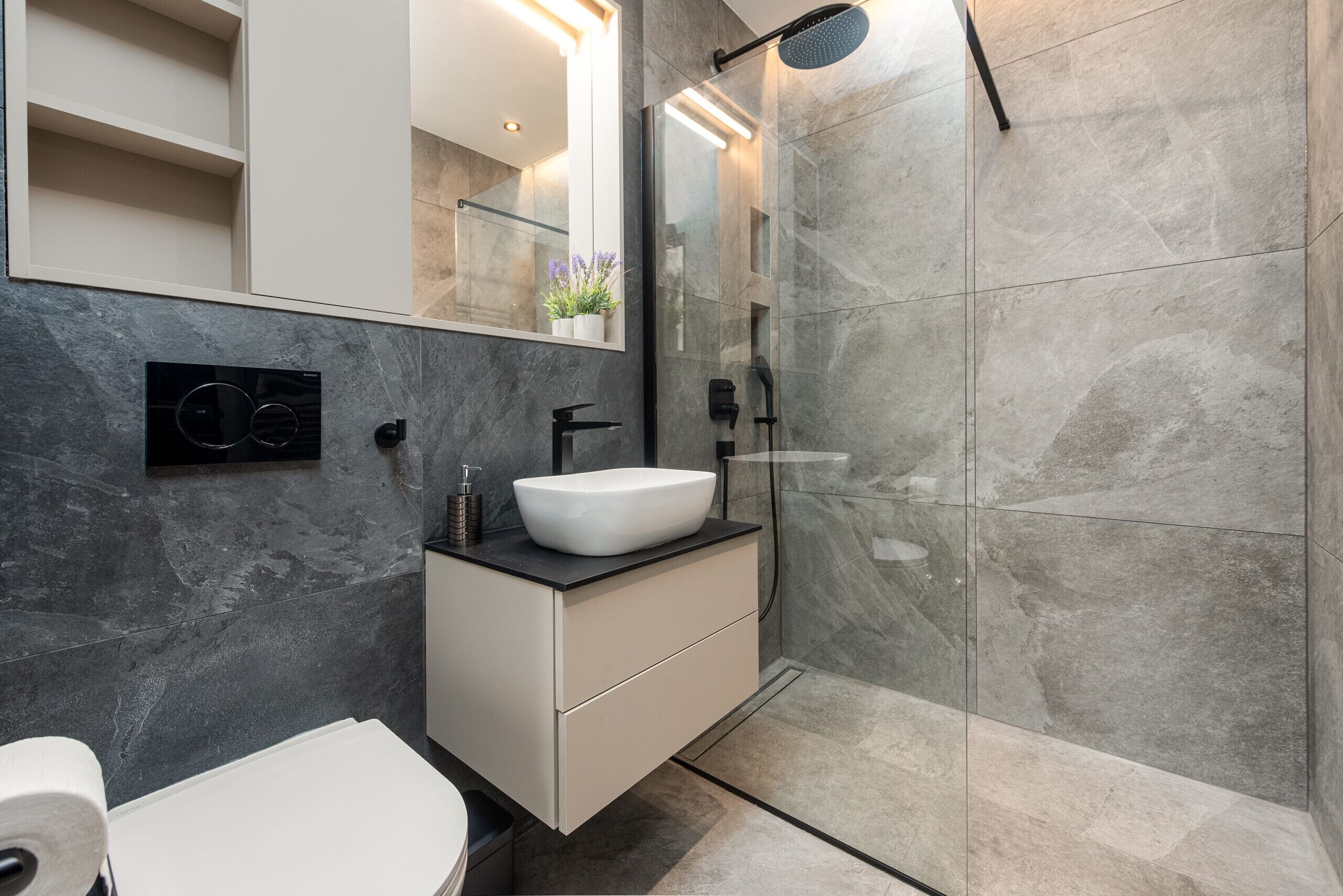 Ein modernes Badezimmer mit einem weißen Waschbecken, einer Duschkabine aus Glas und einer Wand mit Marmorplatten.