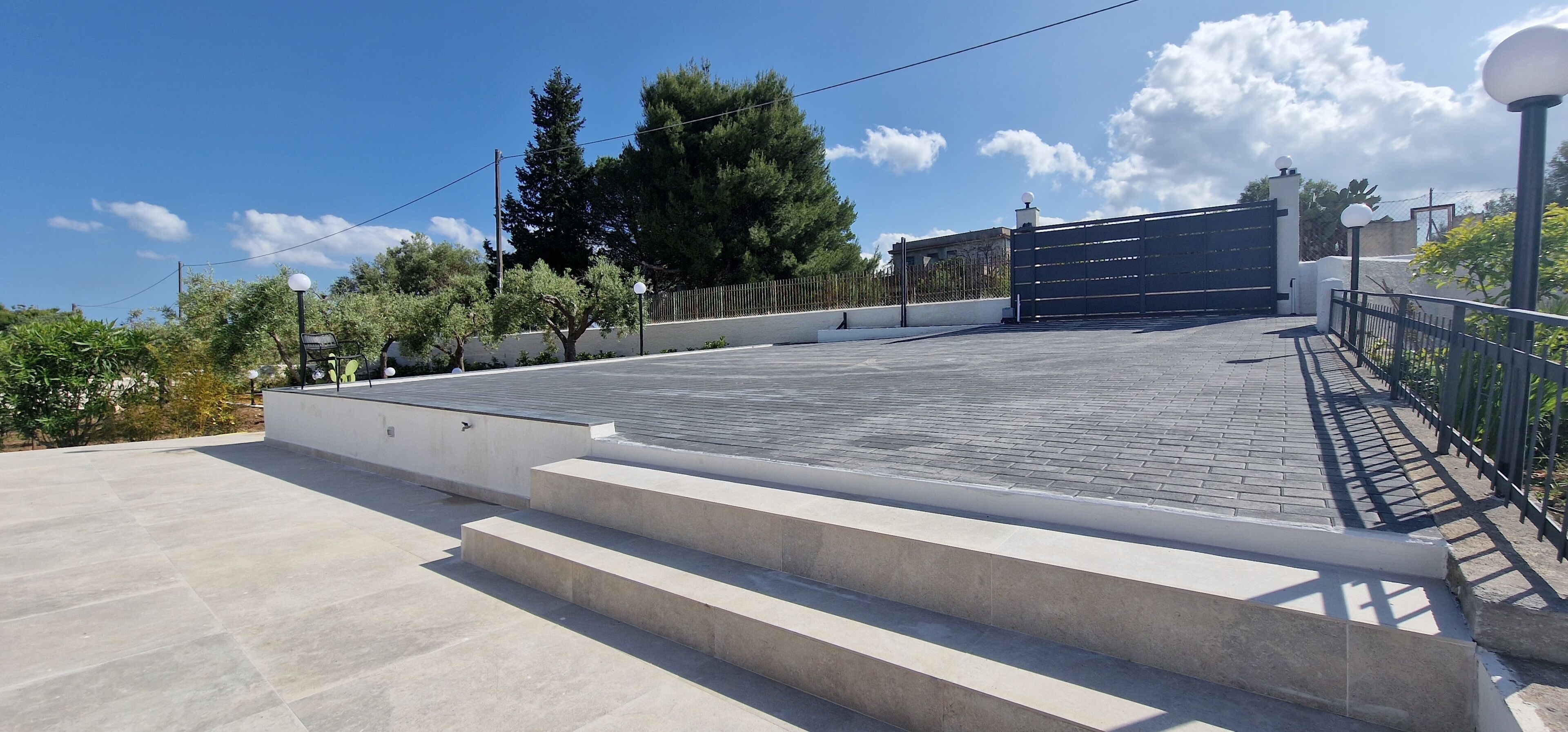 A paved area with steps leading to a gated entrance, surrounded by trees and a clear sky.