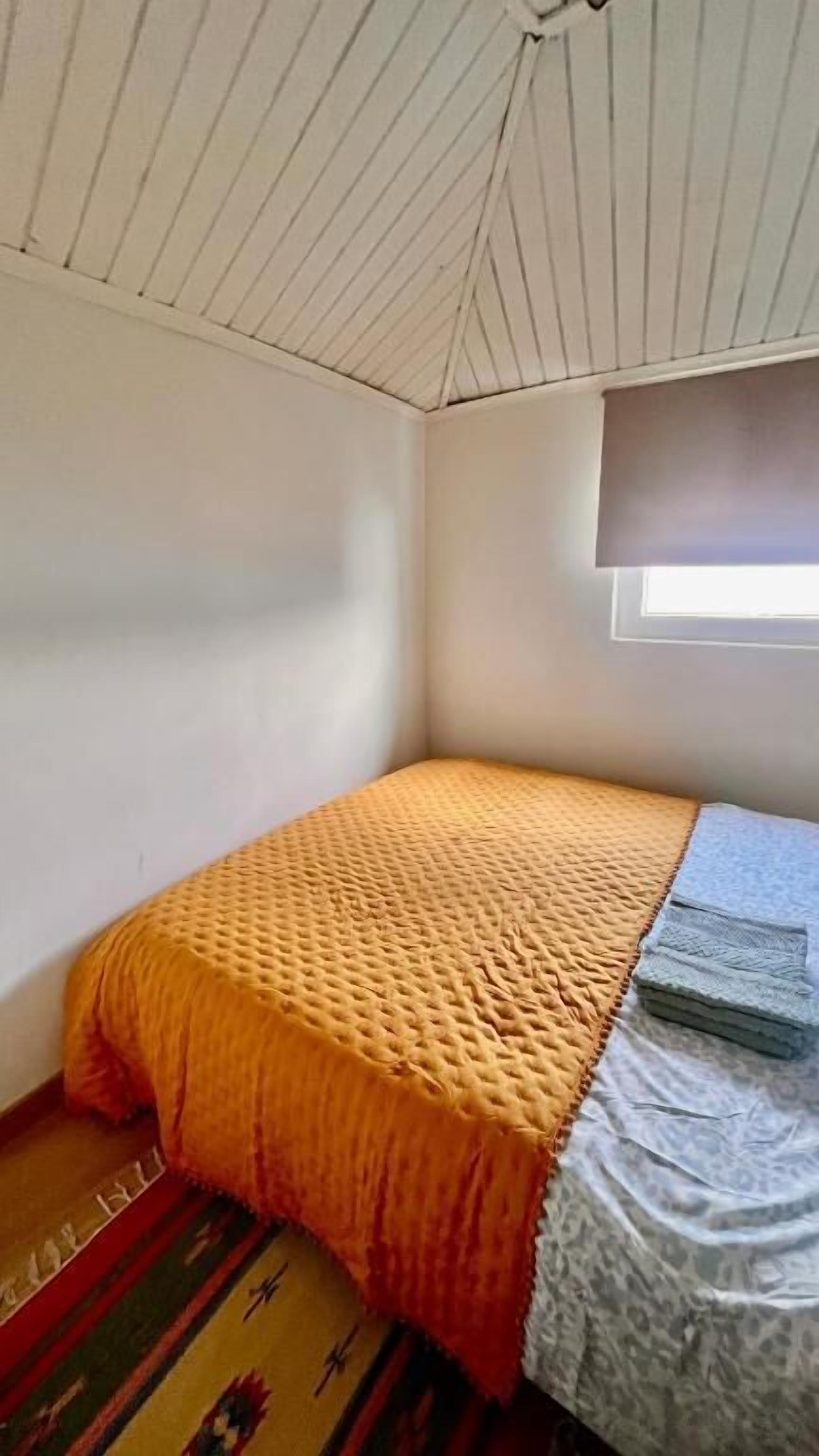 Ein kleines Zimmer mit einem Einzelbett, schrägen Wänden, einem Fenster mit Jalousie und einem gemusterten Teppichboden.