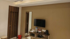 Habitación de hotel con ventilador de techo, televisor de pantalla plana, puerta de madera y una pequeña zona de estar con un taburete.
