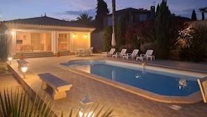 Una piscina con sillas de descanso, un patio con mesa y sillas, y una casa con tejado de baldosas y ventanas grandes.