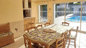 Un comedor con una mesa dispuesta para cuatro, sillas de madera, una cocina con armarios y vistas a una piscina a través de puertas de vidrio.