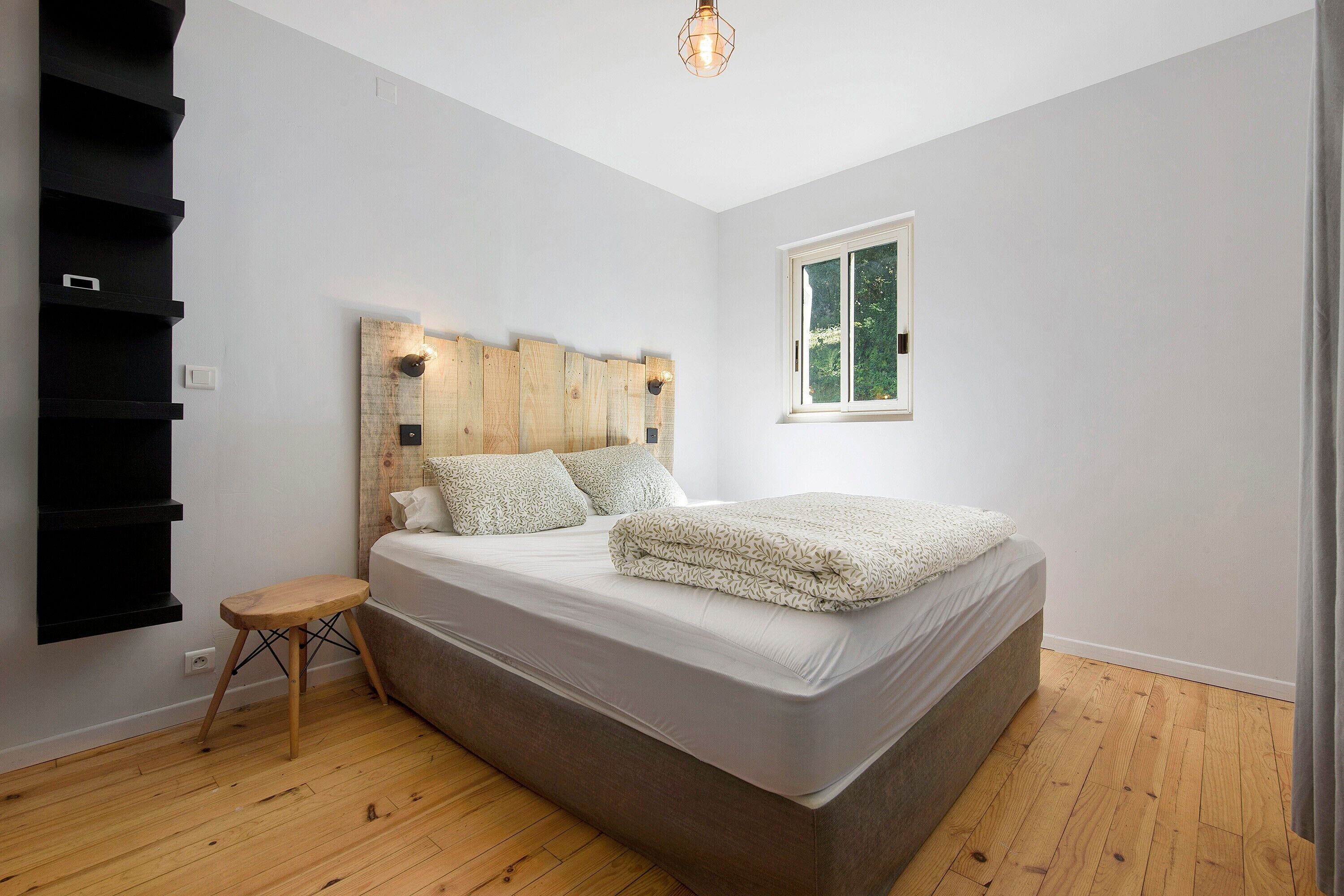 Ein Schlafzimmer mit einem Bett, einem Nachttisch, einem hölzernen Kopfteil, einem Fenster und Holzboden.