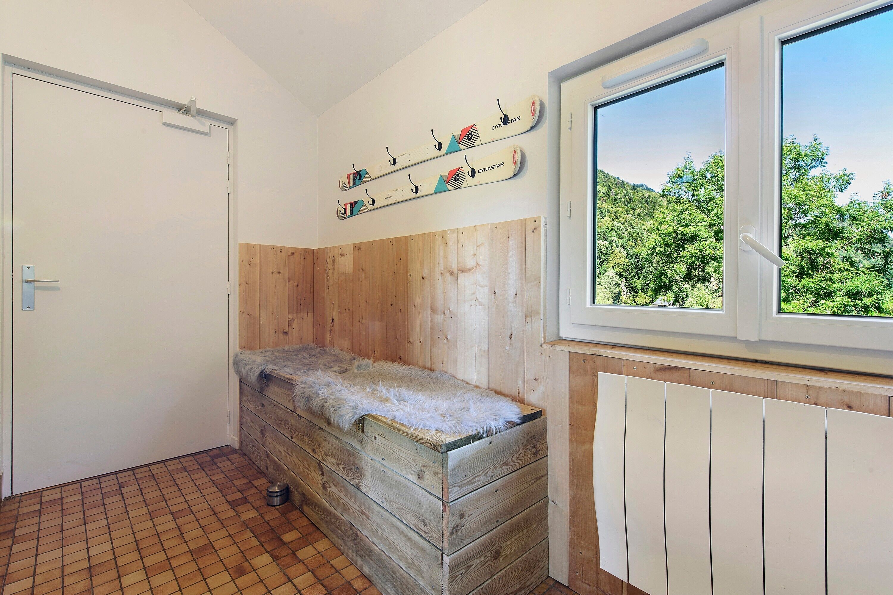 Une pièce avec un banc en bois, une fenêtre donnant sur des arbres et un porte-skis fixé au mur.
