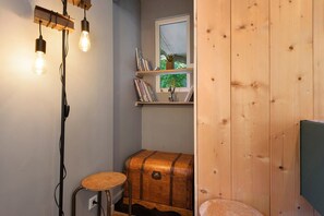 A small, cozy room with a wooden trunk, a stool, and a window with a view of greenery.