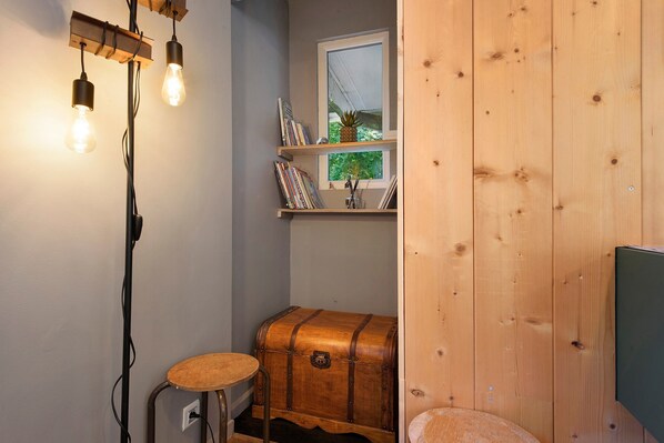 A small, cozy room with a wooden trunk, a stool, and a window with a view of greenery.