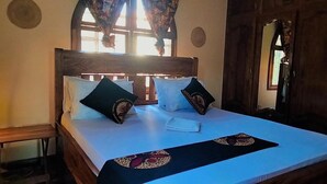 Una cama bien hecha con sábana blanca y dos almohadones decorativos, cabecera de madera y una ventana con cortinas.