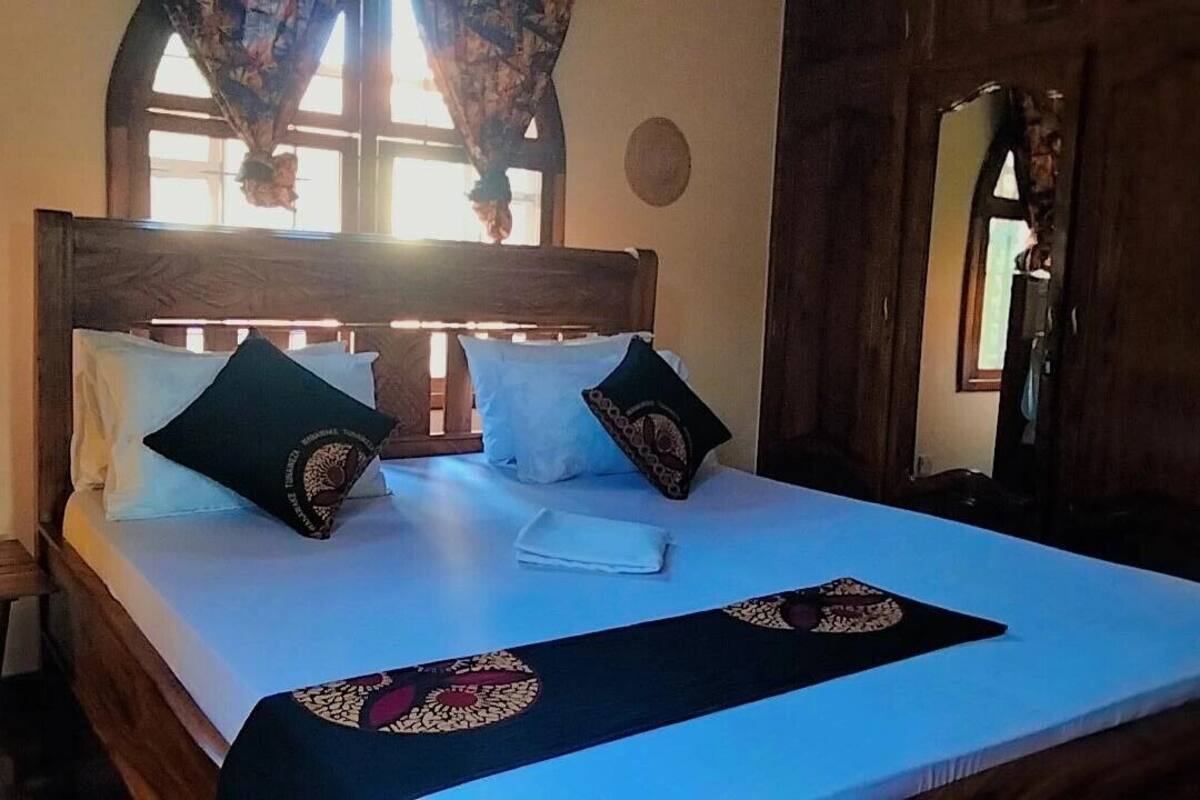 A neatly made bed with a white sheet and two decorative pillows, a wooden headboard, and a window with curtains.