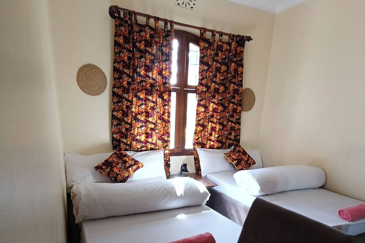 A small room with two beds, a table with water bottles and glasses, and a window with patterned curtains.