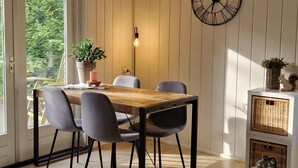 Un coin repas avec une table en bois, des chaises grises, une plante en pot, une horloge murale et un meuble d’exposition en osier.