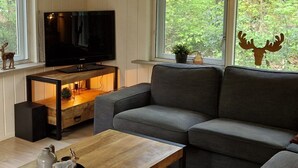 Un salon moderne avec un canapé gris, une table basse en bois, un téléviseur à écran plat posé sur un meuble en bois, et de grandes fenêtres donnant sur une forêt.