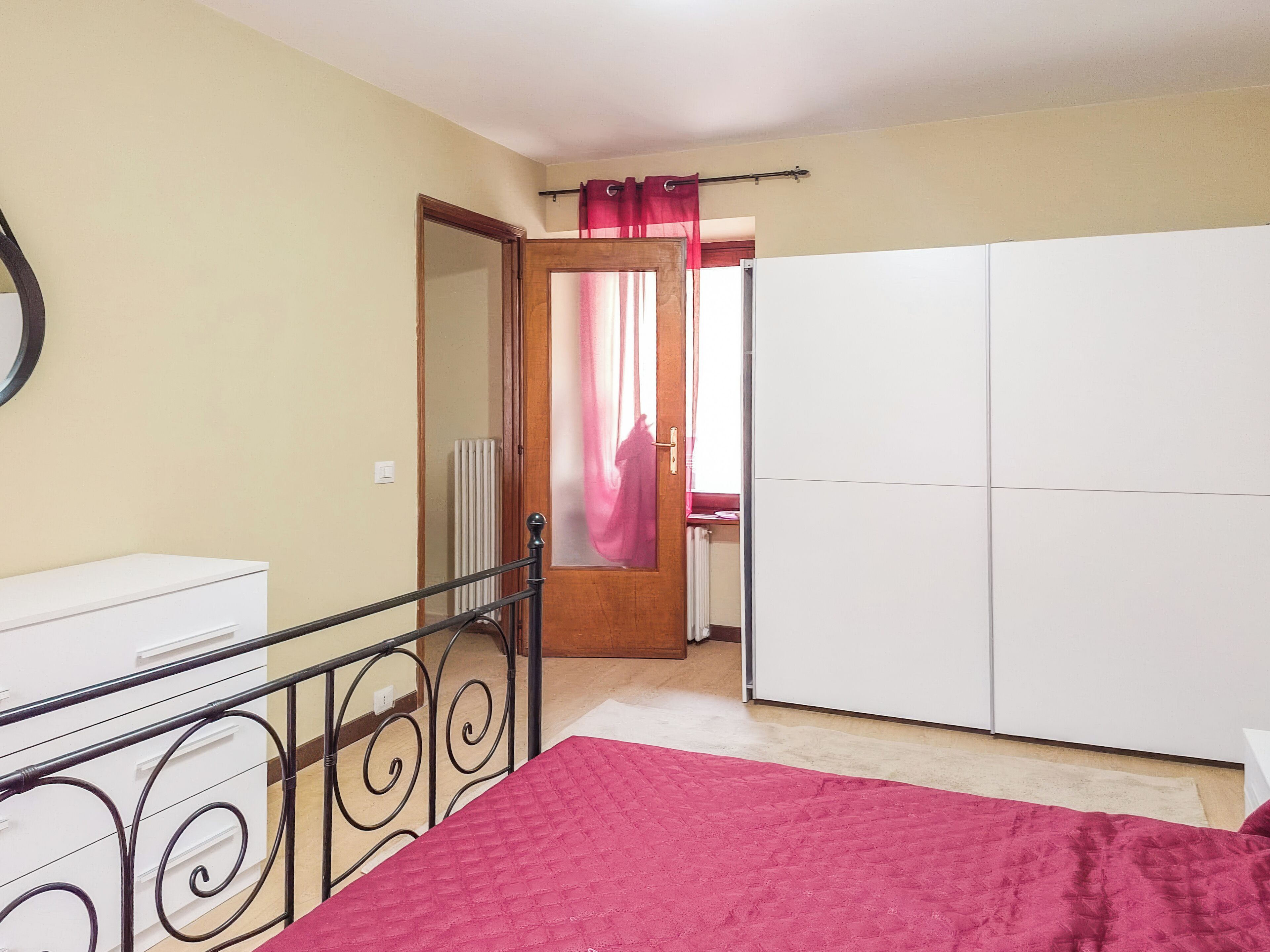 A bedroom with a bed, a wardrobe, a radiator, and a door leading to another room.